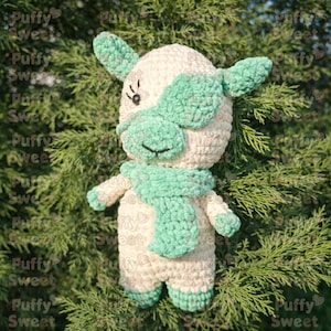May include: A handmade crochet cow doll with a cream-colored body and mint green accents. The doll has a scarf, ears, and a face mask in mint green. The eyes are black, and the doll is standing in front of a green leafy background.