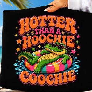 Könnte beinhalten: Ein schwarzes T-Shirt mit einer bunten Grafik eines grünen Alligators mit rosa Sonnenbrille, der auf einem mehrfarbig gestreiften Schwimmreifen im blauen Wasser treibt. Der Text lautet HOTTER THAN A HOOCHIE COOCHIE in fetter, oranger Schrift.