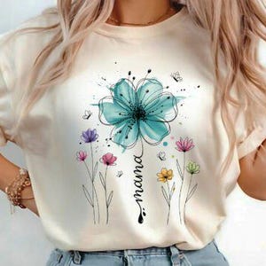 Puede incluir: Camiseta color crema con una gran flor aguamarina en acuarela y la palabra "mama". Flores más pequeñas en rosa, morado y amarillo rodean el diseño principal. Mariposas y salpicaduras de tinta añaden detalles al gráfico floral.