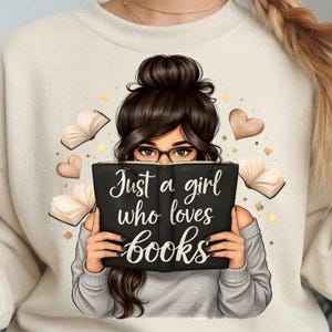 Könnte beinhalten: Ein cremefarbenes Sweatshirt mit einer Grafik einer Person mit dunklem Haar und Brille, die ein schwarzes Buch hält. Das Buch trägt die Aufschrift "Just a girl who loves books" in weißer Schrift. Dekorative Herzen und Bücher sind um die Figur verstreut.