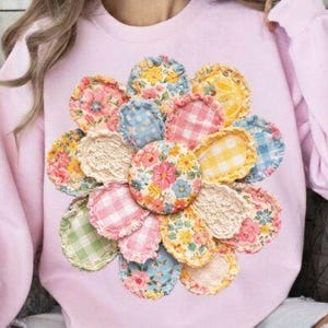Könnte beinhalten: Ein hellrosa Sweatshirt mit einem großen Blumendesign aus verschiedenen Stoffflicken. Die Flicken weisen Blumen-, Vichy- und Spitzmuster in Pastellfarben auf. Das Design ist auf der Brust zentriert.