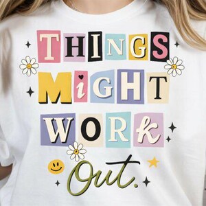 Puede incluir: Camiseta blanca con la frase "THINGS MIGHT WORK OUT" en letras de bloque coloridas. El diseño incluye margaritas, estrellas y una cara sonriente. Una camiseta gráfica casual y optimista.