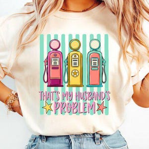 Pode incluir: Camiseta creme com um gráfico de três bombas de gasolina vintage em rosa, amarelo e coral, sobre um fundo listrado turquesa e branco. O texto "THAT'S MY HUSBAND'S PROBLEM" está abaixo, com estrelas.