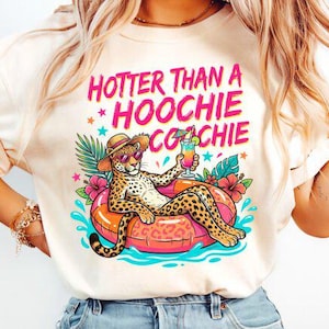 Könnte beinhalten: Cremefarbenes T-Shirt mit dem Text "HOTTER THAN A HOOCHIE COOCHIE" in Pink und einer Grafik eines Leoparden mit Sonnenbrille, der sich auf einem Schwimmring mit einem Cocktail entspannt. Der Leopard ist von tropischen Blumen und Wasser umgeben.