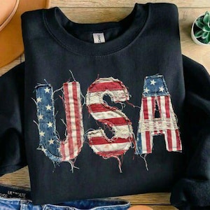 Könnte beinhalten: Schwarzes Sweatshirt mit Rundhalsausschnitt und dem Wort "USA" auf der Vorderseite. Die Buchstaben bestehen aus abgenutztem Stoff im Stil der amerikanischen Flagge. Ein Hut, Jeans und eine kleine Pflanze sind im Hintergrund.