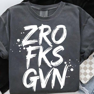 Può includere: T-shirt grigio scuro con la scritta bianca "ZRO FKS GVN" in un font invecchiato. La maglietta è piegata, con una custodia per telefono con motivo a scacchi visibile. Lo sfondo include jeans e un capo nero.