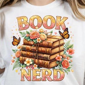 Könnte beinhalten: Weißes T-Shirt mit einem farbenfrohen Grafikdesign. Das Design zeigt die Worte "BOOK NERD" in einer glitzernden Schriftart, mit einem Stapel Bücher, Blumen und Schmetterlingen. Die Bücher sind mit einem Seil zusammengebunden.