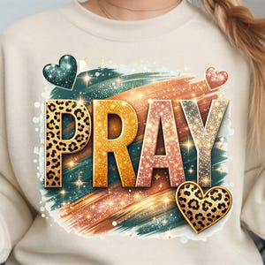 Könnte beinhalten: Ein cremefarbenes Sweatshirt mit einem Grafikdesign, das das Wort PRAY in großen, texturierten Buchstaben zeigt. Die Buchstaben haben Leopardenmuster, Goldglitter und Roségoldglitter. Das Design enthält türkisfarbene und orangefarbene Pinselstriche.