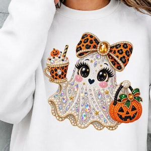 Könnte beinhalten: Ein weißes Langarm-Sweatshirt mit einer Grafik eines Geistes, der mit schillernden Strasssteinen verziert ist. Der Geist trägt eine Schleife mit Leopardenmuster, hält einen Becher mit Leopardenmuster und Kürbisaufsatz und trägt einen orangefarbenen Kürbiseimer.