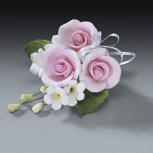 May include: A floral arrangement featuring three pink roses, white blossoms, and green leaves. The roses have a delicate, layered petal design. White ribbon accents the arrangement, which is set against a gray background.