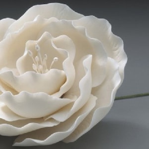 Könnte beinhalten: Eine große, elfenbeinfarbene Kunstblume mit mehreren Schichten zarter Blütenblätter. Die Blume hat ein detailliertes Zentrum und einen grünen Stiel, vor einem grauen Hintergrund. Dieses dekorative Blumenelement ist ideal für Bastelarbeiten oder die Inneneinrichtung.