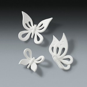 Puede incluir: Tres decoraciones de mariposas blancas con intrincados diseños en las alas. Las mariposas, de diferentes tamaños y formas, presentan diseños calados y superficies texturizadas. Se presentan sobre un fondo gris, creando una exhibición sencilla y elegante.