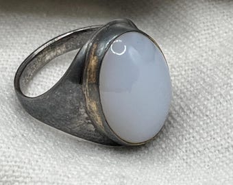 May include: A silver-toned ring with an oval, pale blue gemstone. The ring has a tarnished appearance, with a gold-colored band around the gemstone. The gemstone is smooth and polished, set in a simple, classic design.