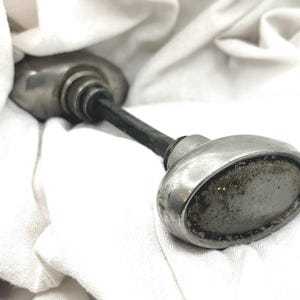 May include: An antique silver-toned door knob with an oval handle and a dark, textured stem. The knob shows signs of age and wear, resting on a white cloth.