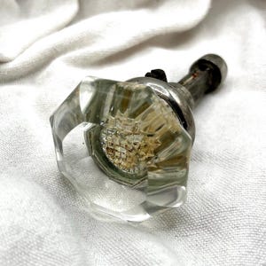 May include: Clear, faceted glass door knob with a silver metal base and stem. The knob has an octagonal shape and a textured interior. The metal stem has a small, round end. The knob is resting on a white, textured surface.