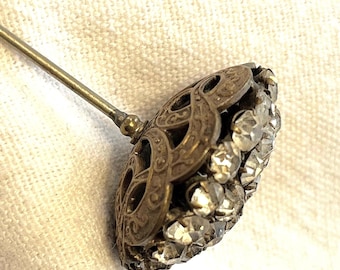 May include: A hat pin featuring a circular, dome-shaped head with an intricate, openwork brass-toned metal frame. The surface is densely set with numerous small, clear, faceted crystals that catch the light, attached to a long, slender metal pin.