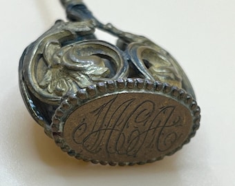 May include: A vintage silver-toned hat pin with an ornate design and the initials "M.H." engraved on the top.