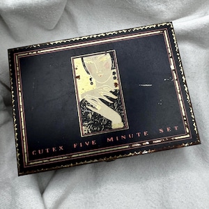 May include: Vintage Cutex Five Minute Set box. The rectangular box is dark blue with gold trim and lettering. The center features a gold and cream illustration of a woman with long earrings and a ring. The box is slightly worn.