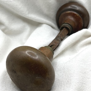 May include: A vintage door knob set featuring two round, dark brown, weathered metal knobs connected by a central square metal spindle. The hardware shows signs of age, including surface scratches and a dull, tarnished patina throughout.