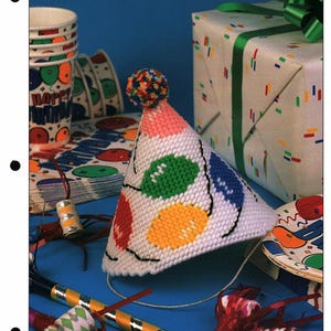 May include: A white party hat with colorful, oval-shaped designs in green, yellow, red, and blue. The hat has a pink top and a multi-colored pom-pom. Other party supplies include cups, plates, and a wrapped gift.