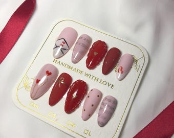 May include: Set of ten false nails with various Valentine's Day themed designs. The nails are in shades of pink and red with gold accents. The designs include hearts, letters, and polka dots. The nails are arranged on a white card with the text "Handmade with Love".