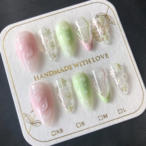 Puede incluir: Un conjunto de diez uñas postizas con un diseño floral. Las uñas son de varios tamaños, etiquetados como XS, S, M y L. Las uñas están pintadas en tonos de rosa y verde con flores blancas y un fondo verde claro. El texto "HANDMADE WITH LOVE" está impreso en la tarjeta.