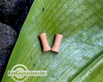 May include: Two wooden ear gauges, one brown and one slightly lighter brown, are displayed on a green leaf. The gauges are cylindrical and have a slightly flared opening at each end. The text "EarGaugesJewelry" is visible in the lower left corner of the image.