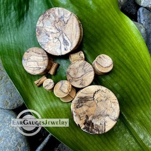 May include: A set of eight wooden ear gauges in various sizes, featuring a unique wood grain pattern with brown and beige tones. The gauges are displayed on a green leaf.