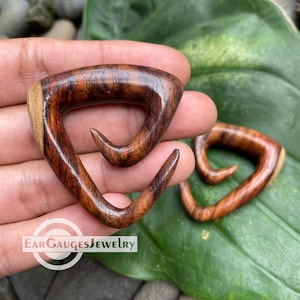 May include: Two wooden ear gauges with a triangular shape and a curved hook design. The gauges are made of dark brown wood with lighter brown grain patterns. The text "Ear Gauges Jewelry" is visible.