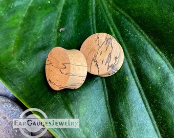 May include: Two wooden ear gauges with a natural wood grain pattern and black wood burning designs. The gauges are on a green leaf.