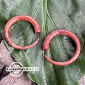 May include: A pair of reddish-brown wooden ear gauges, spiral-shaped, displayed on a large green leaf. The gauges have a polished finish. The text "Ear Gauges Jewelry" is visible at the bottom.