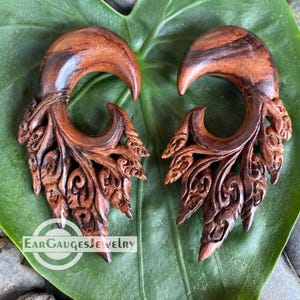 May include: Two carved wooden ear gauges with detailed leaf and scroll patterns. The gauges are a deep brown hue and have a curved, hook-like form. The text "Ear Gauges Jewelry" is visible.