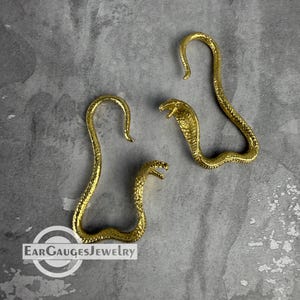 May include: A pair of gold-toned snake-shaped ear gauges with a textured, scale-like surface. Each piece features a curved body forming a hook shape with a detailed cobra head at the end. The text EARGAUGESJEWELRY is visible in the bottom corner.