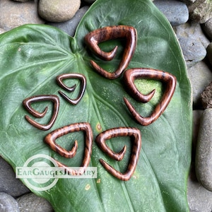 May include: A collection of wooden ear gauges in various sizes and shapes, featuring a triangular design with a curved hook. The gauges are a rich brown color with visible wood grain, set against a green leaf.