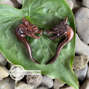 May include: Two wooden ear gauges shaped like dragon heads. The gauges are dark brown and have intricate carvings. They are displayed on a green leaf.