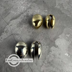 May include: Two pairs of polished gold-tone teardrop-shaped ear weights with a smooth, reflective surface and a vertical opening. The text EARGAUGESJEWELRY is visible in the lower left corner of the image, featuring a circular logo design.