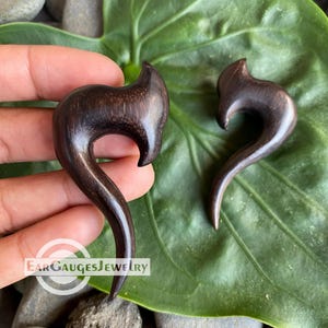 May include: A pair of dark brown wooden ear gauges shaped like a curved hook. The gauges are shown against a green leaf background.