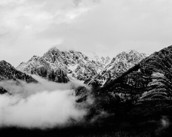 May include: A black and white photograph of a mountain range covered in snow and clouds. The mountains are in the background, and the clouds are in the foreground. The image is taken from a low angle, so the mountains appear to be very tall.