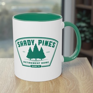 May include: White ceramic mug with a green interior, handle, and a green logo that reads "SHADY PINES RETIREMENT HOME" with a tree graphic. The mug is on a wooden surface.