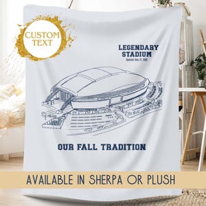 May include: White blanket featuring a blue line drawing of a stadium. Text includes "LEGENDARY STADIUM" and "OUR FALL TRADITION." The blanket also has a gold circle with the words "CUSTOM TEXT" and the text "AVAILABLE IN SHERPA OR PLUSH."