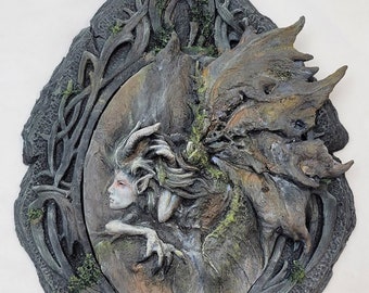 May include: A gray, sculpted wall hanging featuring a female figure with horns and wings, surrounded by a leafy, vine-like frame. The figure is partially obscured by the frame, and the artist's signature, "Sprite Hansen", is visible at the bottom.