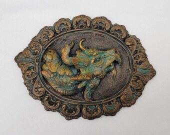 May include: A green and gold dragon sculpture in a decorative oval frame. The dragon is depicted in a fierce pose with its mouth open and teeth bared. The frame is made of metal and has a distressed finish.