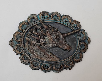May include: A bronze-colored unicorn head sculpture with a single horn, set in an oval frame with ornate gold and teal details.
