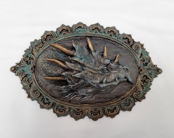May include: A bronze and turquoise framed wall sculpture of a dragon's head. The dragon has a textured, scaly skin and sharp horns. The frame is ornate and has a distressed finish.