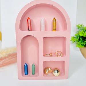 May include: A light pink arched display shelf with multiple compartments containing six small crystal points in red, clear, and brown, two gold-toned rings, and two gold-toned spherical earrings arranged on the various shelves.