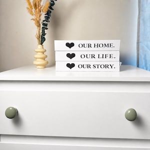 May include: A stack of three white rectangular blocks with black text and heart icons. The top block reads OUR HOME., the middle block reads OUR LIFE., and the bottom block reads OUR STORY. Next to the blocks is a beige vase with dried plants.