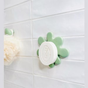Pode incluir: Um saboneteira verde claro com uma barra de sabão branca. A saboneteira tem a forma de uma flor estilizada e está montada numa parede de azulejos brancos. O texto no topo diz "STOP WASTING SOAP" e na parte inferior "DRIES FASTER. NO MESS".
