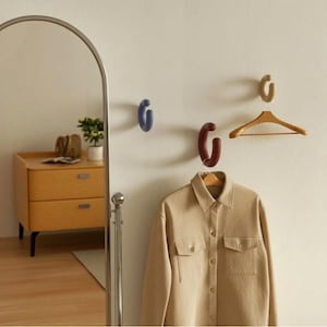 May include: A neutral-toned room features a wooden dresser, a full-length mirror, and a beige button-up shirt hanging on a wooden hanger. Three curved wall hooks in blue, burgundy, and beige add a pop of color to the white wall.