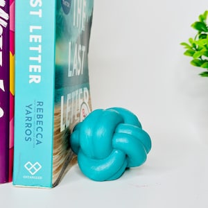 Könnte beinhalten: Ein türkisfarbener dekorativer Knoten liegt neben einem Buch. Der Buchrücken zeigt die Titel "LAST LETTER" und "THE LAST LETTER" von Rebecca Yarros. Der Knoten ist eine glatte, solide Skulptur, die einen Farbtupfer hinzufügt.