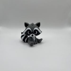 May include: A gray and white raccoon figurine sitting on a white surface. The raccoon has a black mask around its eyes, a striped tail, and is in a seated position. The figurine is a cute, cartoon-like representation of the animal.